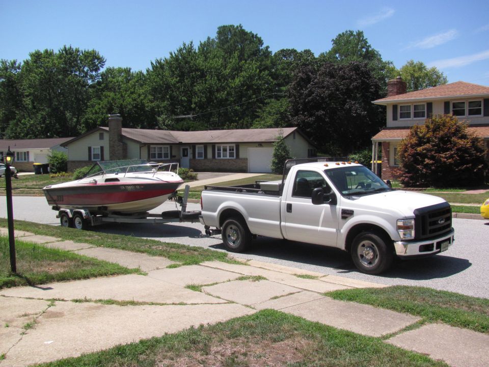 White Ford Super Duty pickup towing a red-and-white boat on a trailer down a residential street