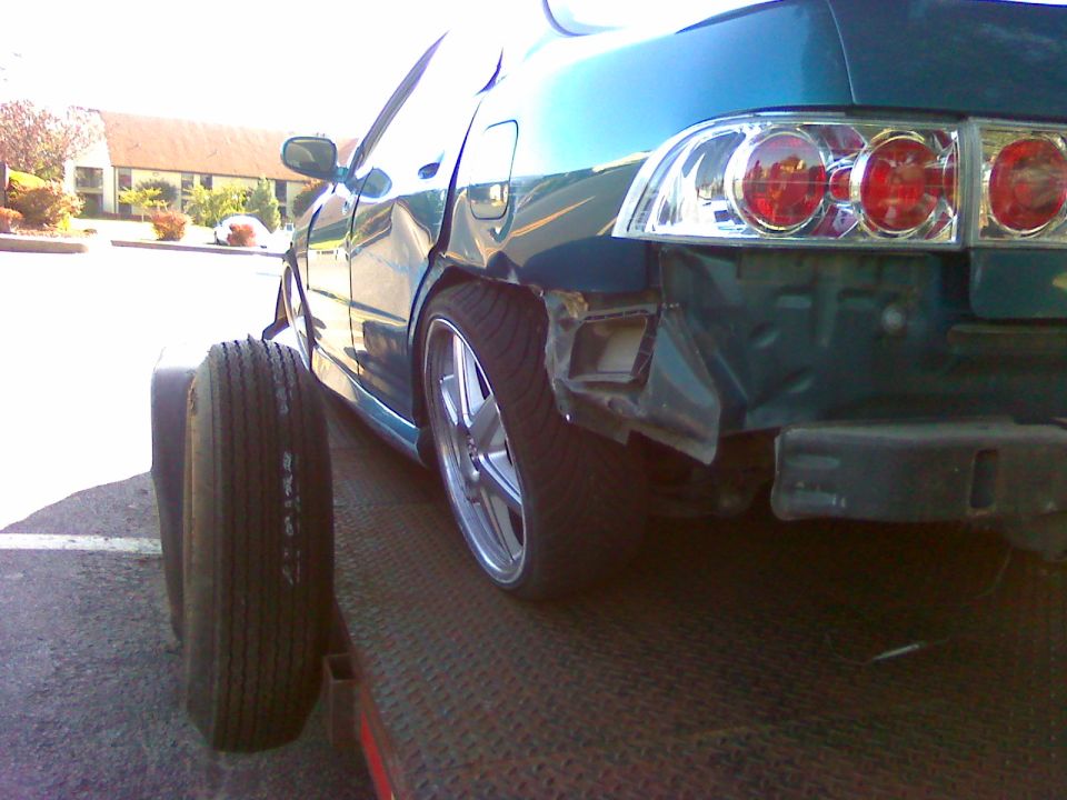 Teal sedan with damaged rear bumper and quarter panel on a flatbed tow truck