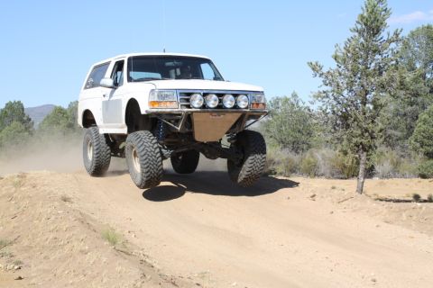 1996 Ford Bronco - A modified white SUV jumps over a dirt ramp, kicking up dust under a clear blue sky.