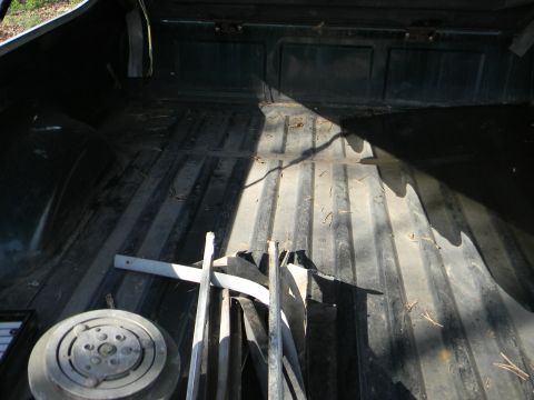 1979 Ford Ranchero - The interior of a truck bed is shown, containing various metal parts and a circular component, with sunlight creating distinct shadows.
