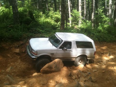 2005 Jeep Wrangler - White Ford Bronco tackles a challenging off-road trail in a wooded area. A club sticker is visible on the windshield.