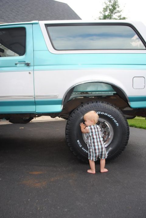 1994 Ford Bronco - A baby inspects the massive tire of a two-tone blue and white SUV.