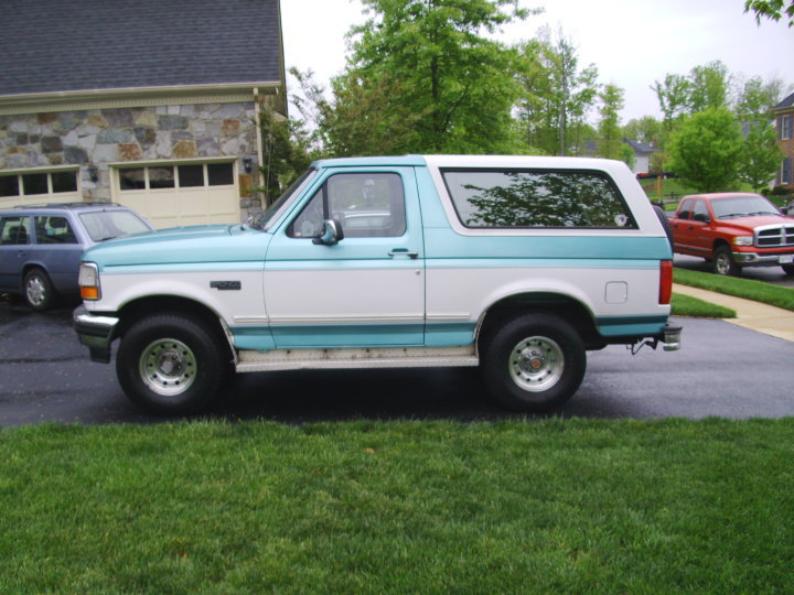 Side view of a two-tone blue and white 1994 Ford Bronco parked on grass next to a house. Another SUV and a red pickup truck are visible