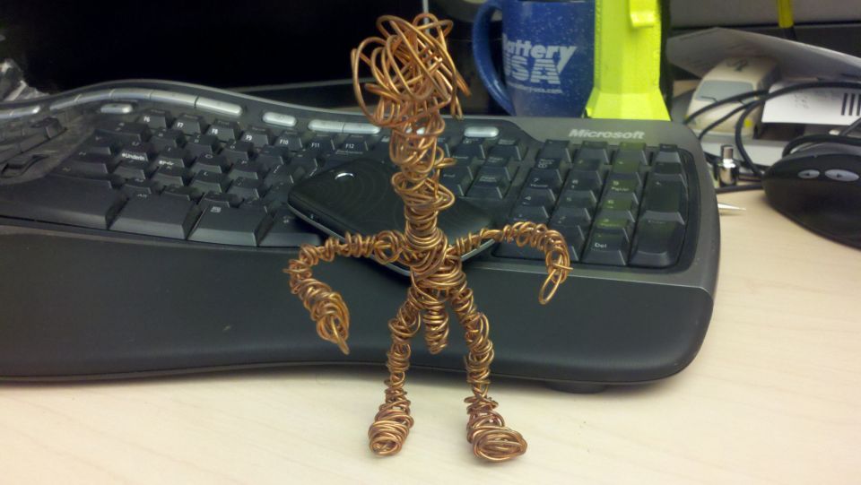 Wire sculpture of a person standing on a desk in front of a keyboard and a mug