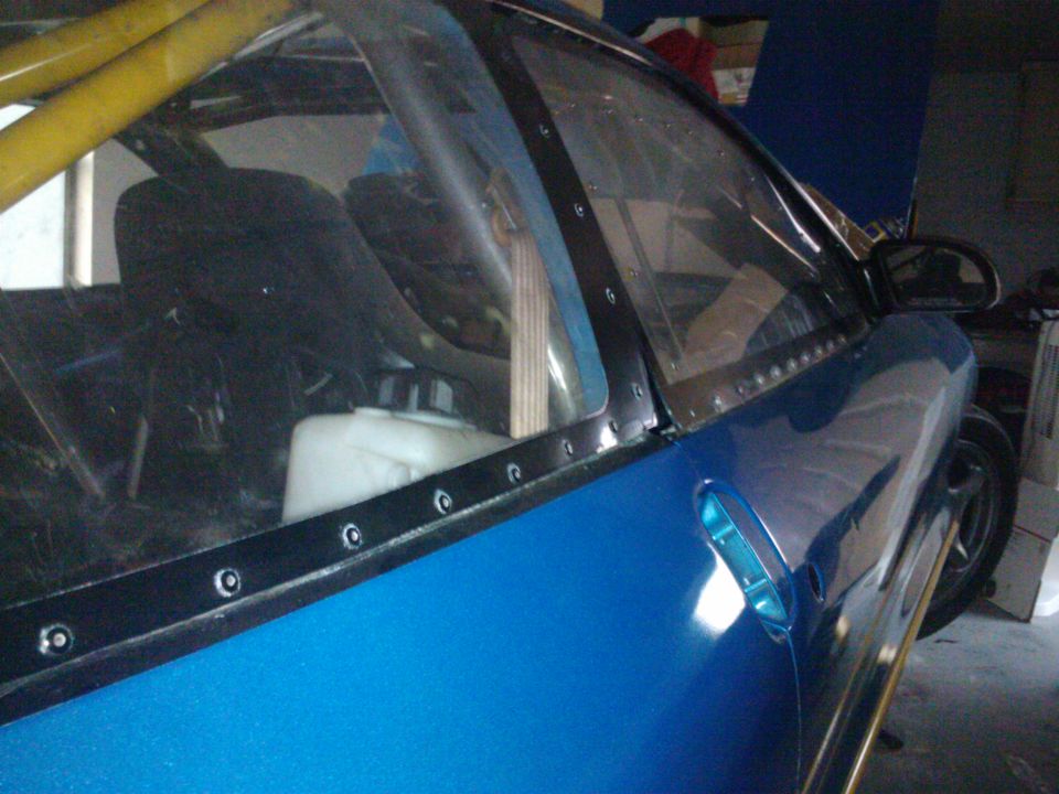 Blue custom car interior with yellow roll cage, clear plastic windows riveted on, and a white fluid tank visible
