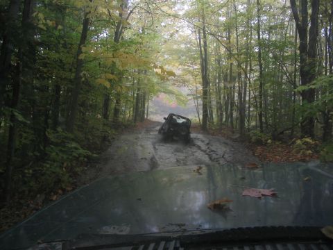 1986 Ford Bronco - A dark SUV navigates a muddy path through a dense forest during autumn.