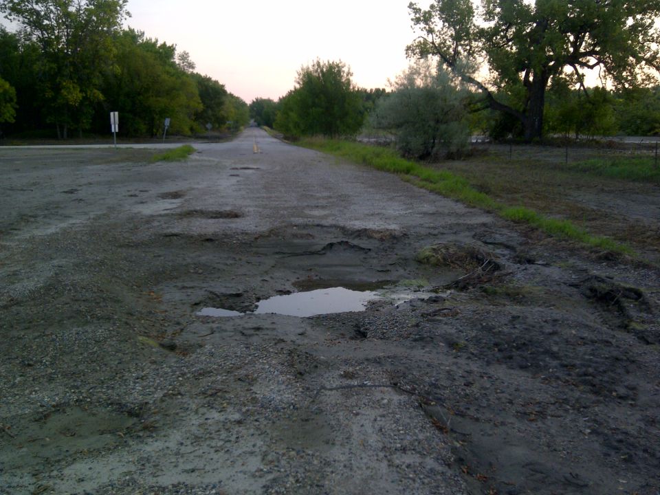 Dirt road with many deep potholes and puddles, lined with trees