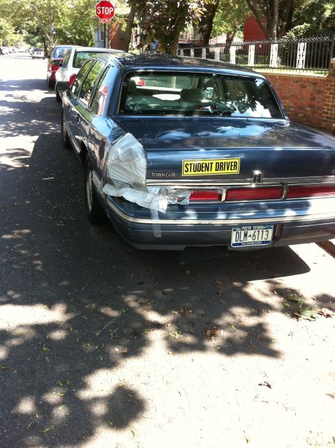 2007 Subaru Impreza - A dark sedan with a "STUDENT DRIVER" sticker and a rear bumper crudely repaired with plastic wrap and tape is parked on a street.
