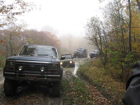 1986 Ford Bronco - A convoy of SUVs navigates a challenging muddy path through an autumn forest.