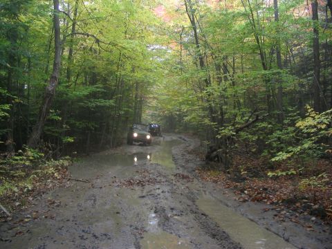 1986 Ford Bronco - Off-road vehicles traverse a challenging muddy path surrounded by lush green trees.
