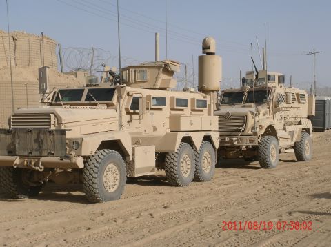1988 Ford Bronco - Two tan military armored vehicles are parked on a dirt road in a desert environment.