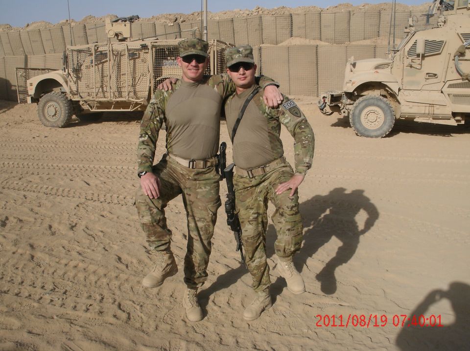 Two soldiers in camouflage uniforms and caps pose in front of military vehicles in a desert environment. One soldier has his arm around the other's shoulder