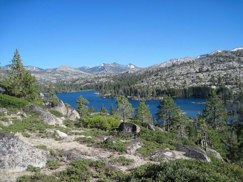 1986 Ford Bronco - A vast mountain landscape unfolds with a tranquil blue lake reflecting the clear sky, bordered by dense pine forests and , rocky hills.