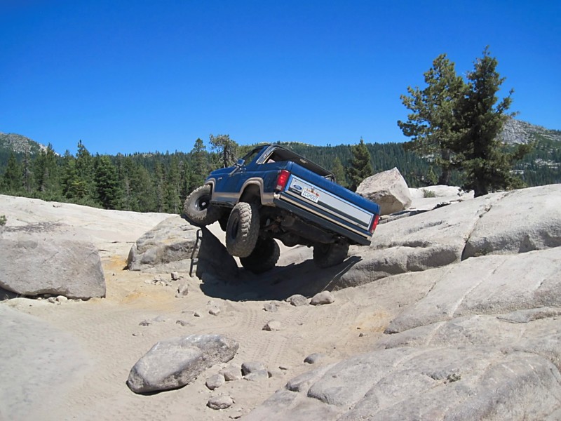 1986 Ford Bronco climbing over large rocks on a dirt trail with trees and mountains in the background