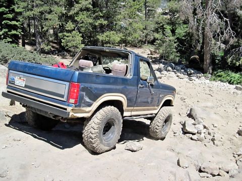 1986 Ford Bronco - A blue Ford Bronco navigates a challenging rocky terrain on a sunny day.