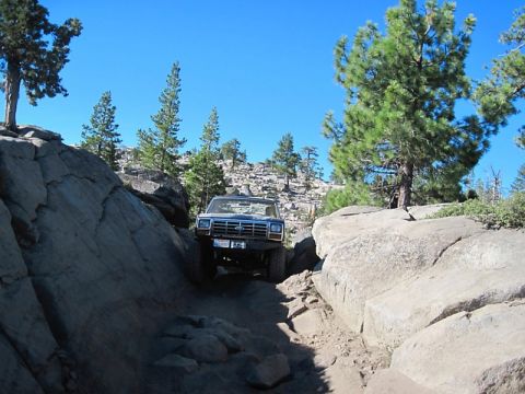 1986 Ford Bronco - A blue SUV carefully maneuvers through a tight, rocky passage on an off-road trail under a clear blue sky.