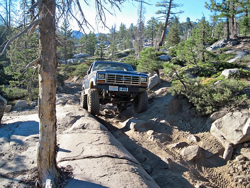 1980 Ford Bronco navigating a steep, rocky off-road trail with large tires and lifted suspension. License plate reads '205001'