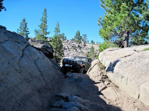 1986 Ford Bronco - Ford Bronco tackling a challenging rocky trail in a mountainous, wooded area under a clear blue sky.