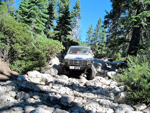 1986 Ford Bronco - This 1980s Ford Bronco is shown off-roading on a challenging, boulder-strewn path through a forest.