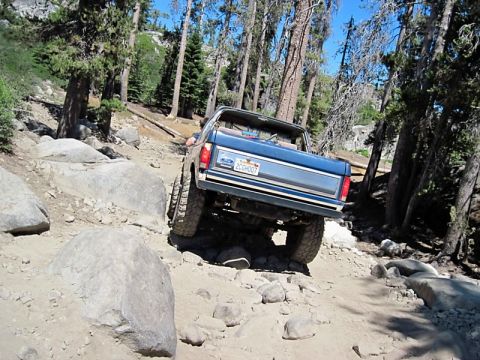 1986 Ford Bronco - 1986 Ford Bronco tackles a challenging off-road trail with large rocks and trees.