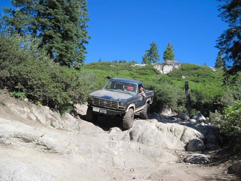 1986 Ford Bronco - A blue Ford Bronco navigates a rocky off-road path under a clear blue sky.