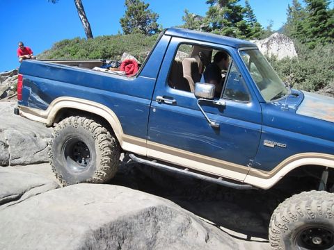 1986 Ford Bronco - A blue Ford Bronco navigates a rocky trail, its tires gripping the stones as it climbs.