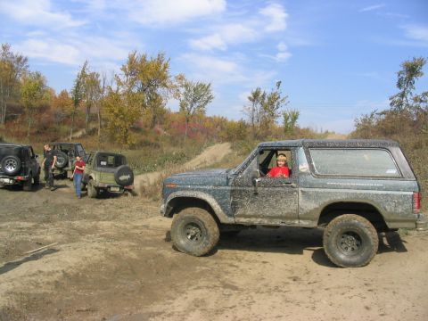 1986 Ford Bronco - A group of off-road vehicles are gathered on a dirt track with trees in the background.
