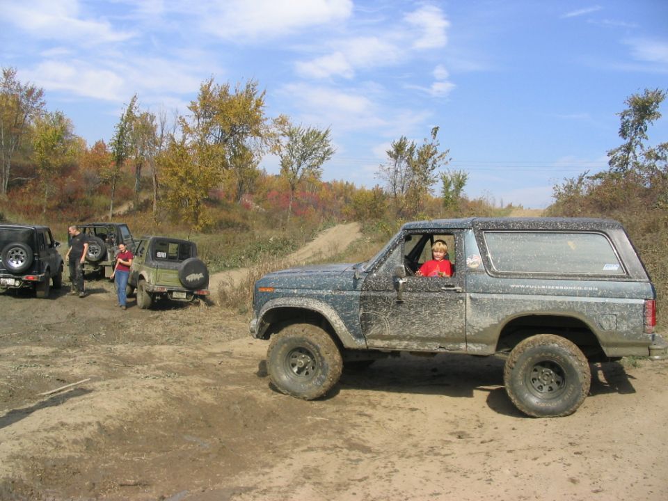 Mud-splattered dark green 1980 Ford Bronco, driver in red shirt, side view, off-road setting