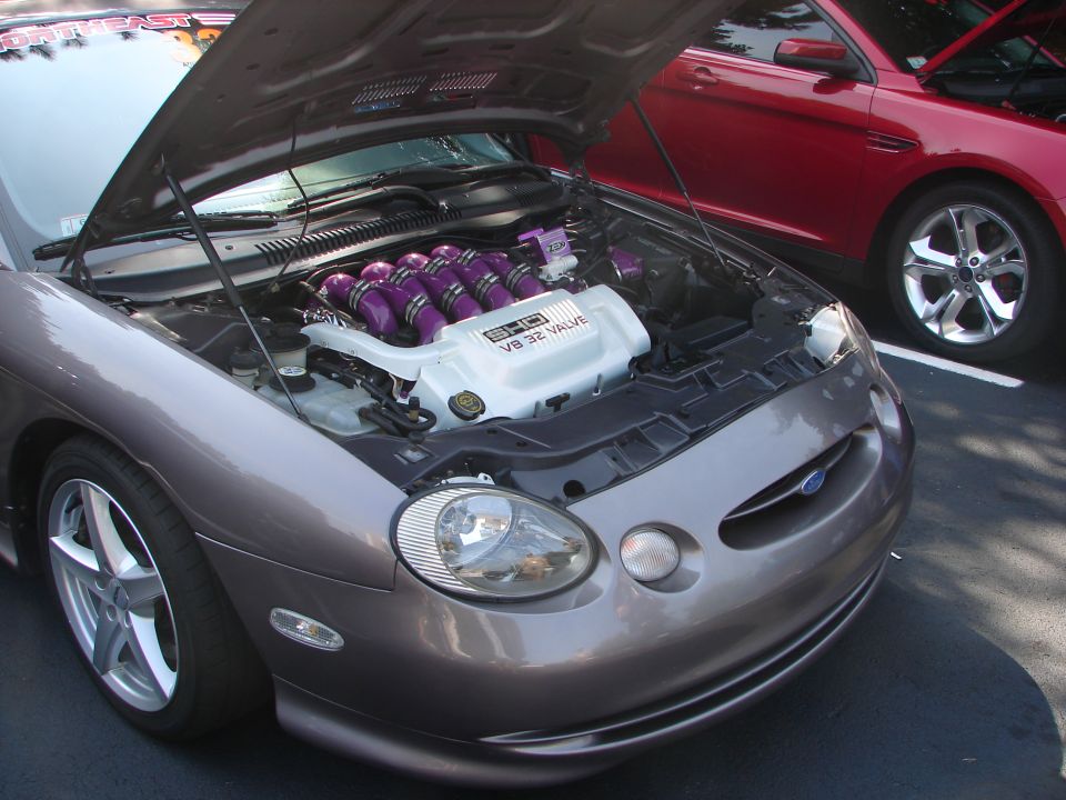 Gray Ford Taurus with hood open, showing a V8 32 valve engine with purple intake runners. A red Ford Taurus is parked behind it