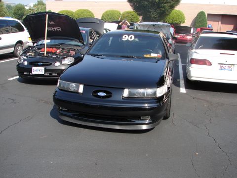 1992 Ford Taurus - A black Ford Taurus with a '90' sticker on its windshield is parked in a lot with other vehicles.