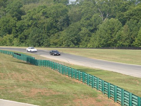 1992 Ford Taurus - Two sedans race on a track bordered by green guardrails and lush trees.