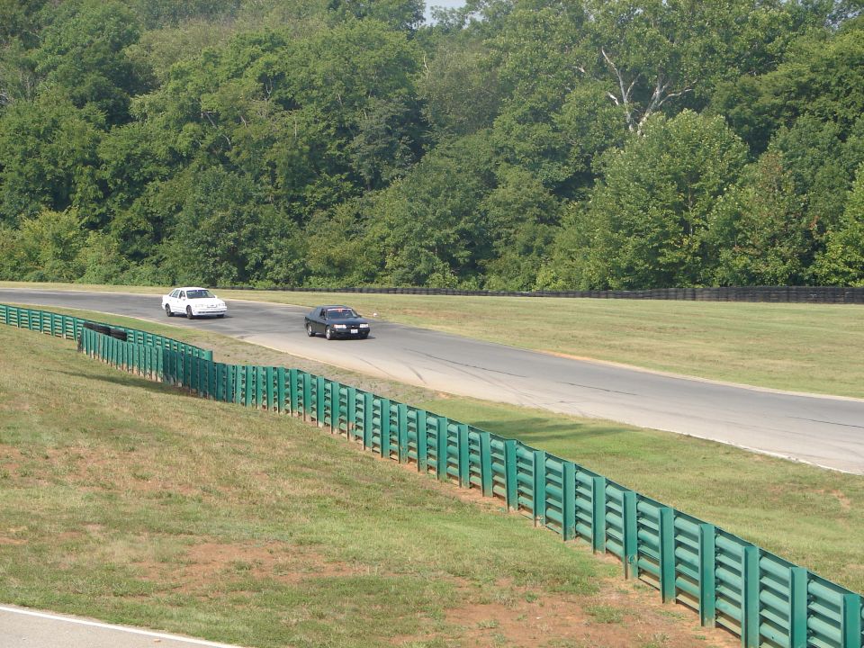 Black sedan and white sedan on a racetrack, with green guardrails and trees in the background