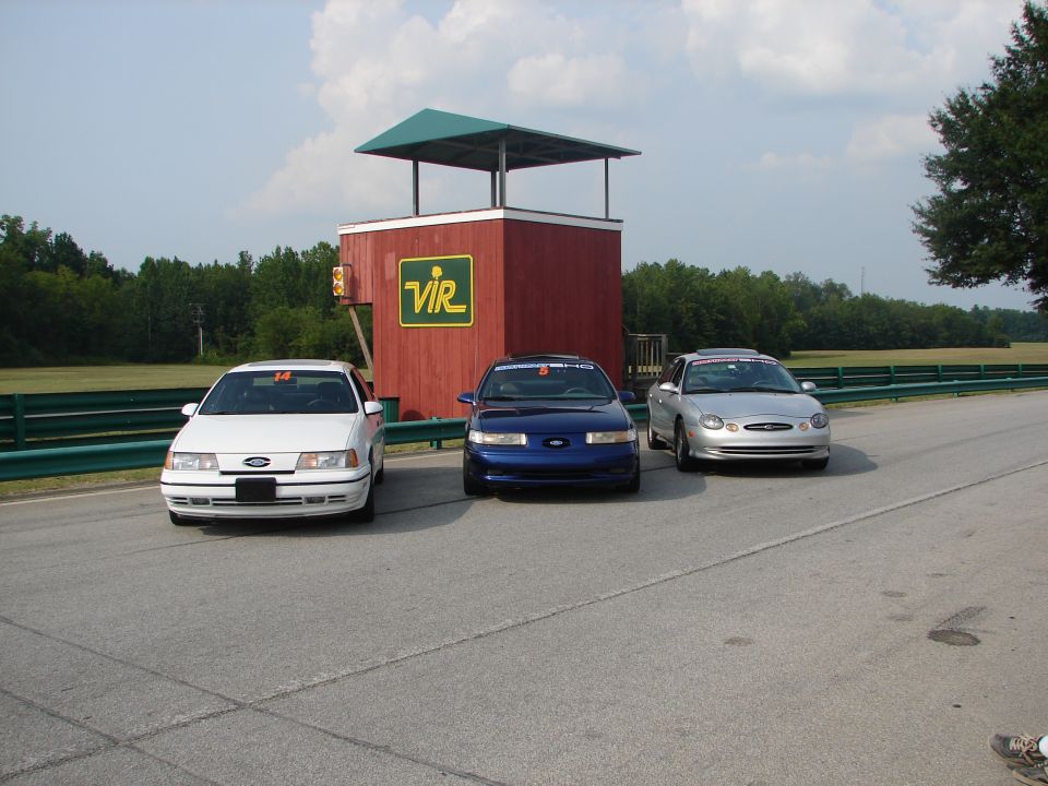 Three Ford Taurus sedans parked on a racetrack in front of a red building with a green roof. The white and blue cars are older models, the silver one appears to be a later generation