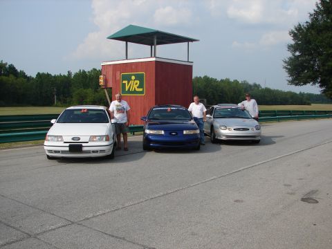 1992 Ford Taurus - Three Ford Taurus sedans are parked on a racetrack, with a group of men standing beside them near a race control tower.