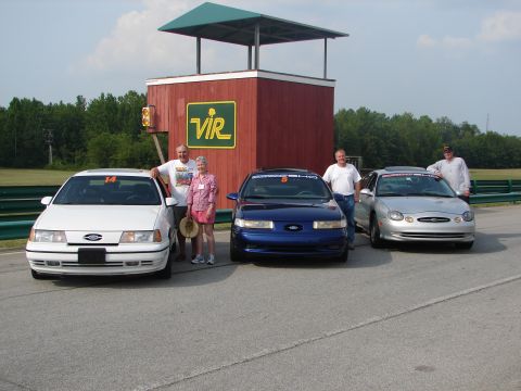 1992 Ford Taurus - Three Ford Taurus sedans are lined up on a race track with people posing in front of a building.