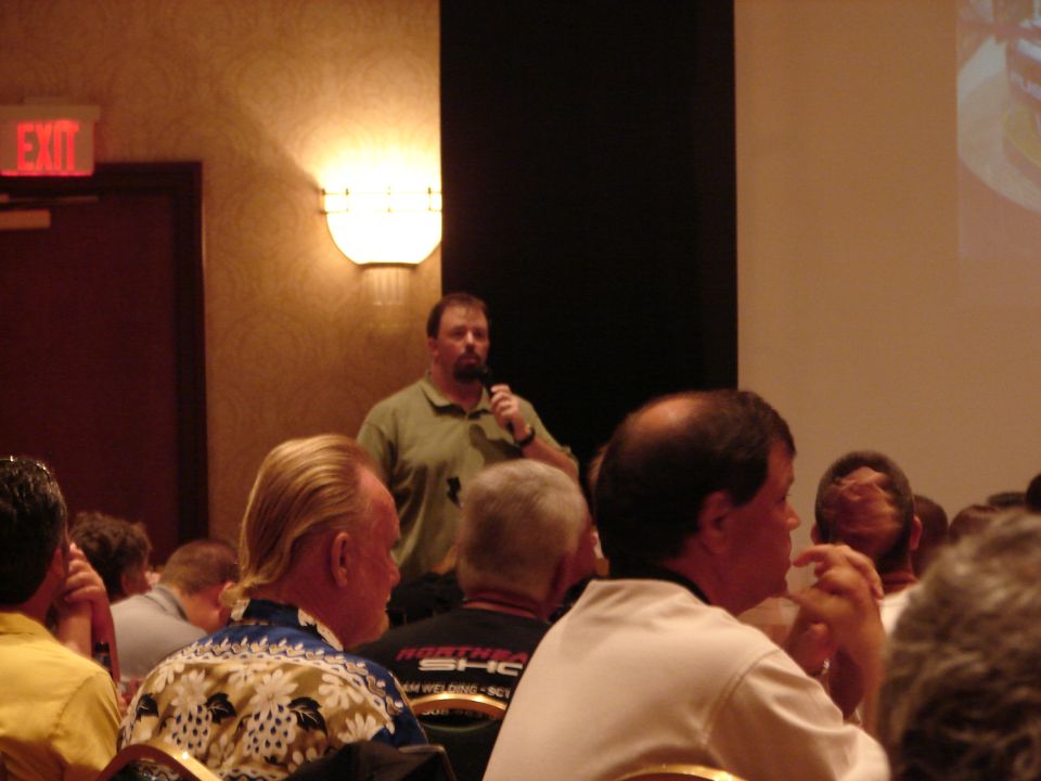 Man speaking into a microphone at a convention, with an audience seated in front of him