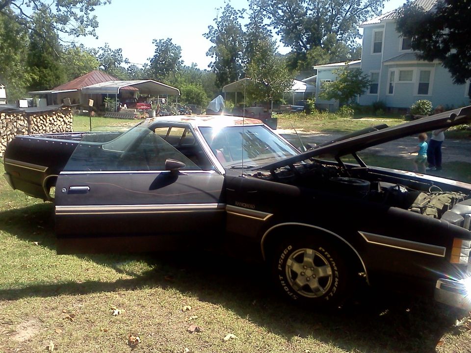 Black Ford Ranchero with hood and driver door open, parked on grass