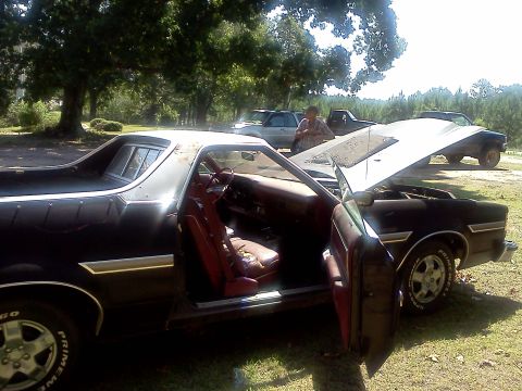 1979 Ford Ranchero - A black 1979 Ford Ranchero with its hood and driver's door open, showing its red interior, sits in a grassy area with other vehicles in the…