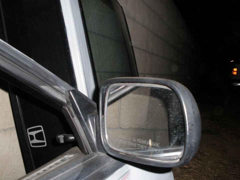 Passenger mirror on white Honda at night, Honda H decal on pillar, metal building behind