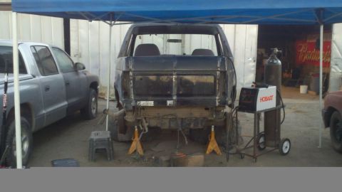 1989 Ford Bronco - A dark SUV body is undergoing repairs, supported by jack stands, with a welding setup and another vehicle present.