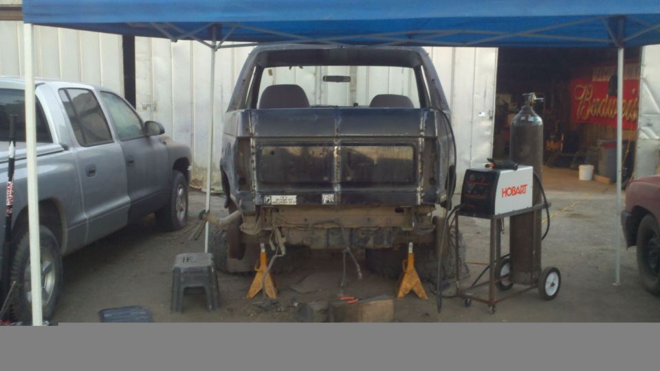 Rear view of a dark SUV body on jack stands, next to a silver pickup truck. Welding equipment is nearby