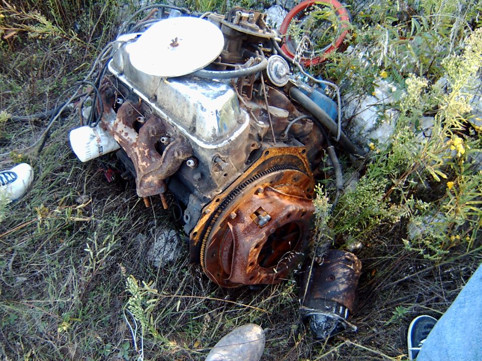 Rusty carbureted V8 engine with chrome air cleaner lying in weeds beside a starter motor