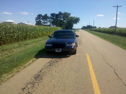 1999 Mercury Grand Marquis - Dark sedan parked on a rural road with a cornfield and silos in the background.