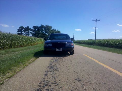 1999 Mercury Grand Marquis - A dark sedan is parked on the side of a rural road with a cornfield on one side and a paved road on the other.