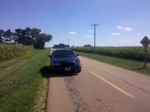 1999 Mercury Grand Marquis - A dark blue sedan is parked on the shoulder of a road bordered by cornfields under a clear blue sky.