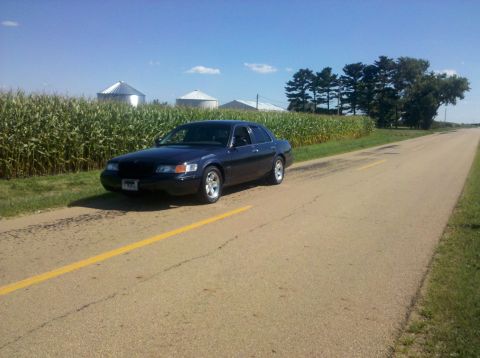 1999 Mercury Grand Marquis - A dark blue 1999 Mercury Grand Marquis sits parked on the side of a country road, with a cornfield and silos in the background.