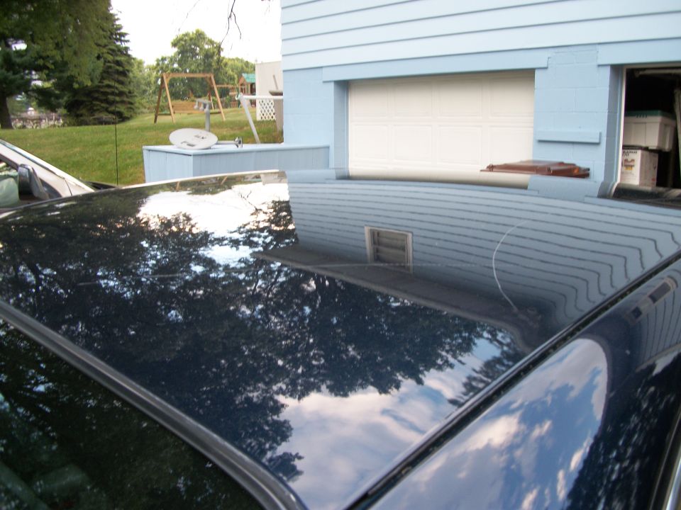 Polished dark blue sedan roof reflecting sky, parked by a light blue house and garage