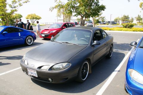 1995 Ford Taurus - A matte black Ford Escort coupe is parked in a lot with other vehicles visible.