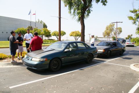 1995 Ford Taurus - A group of people gather around a dark green sedan with a modified hood in a parking lot.