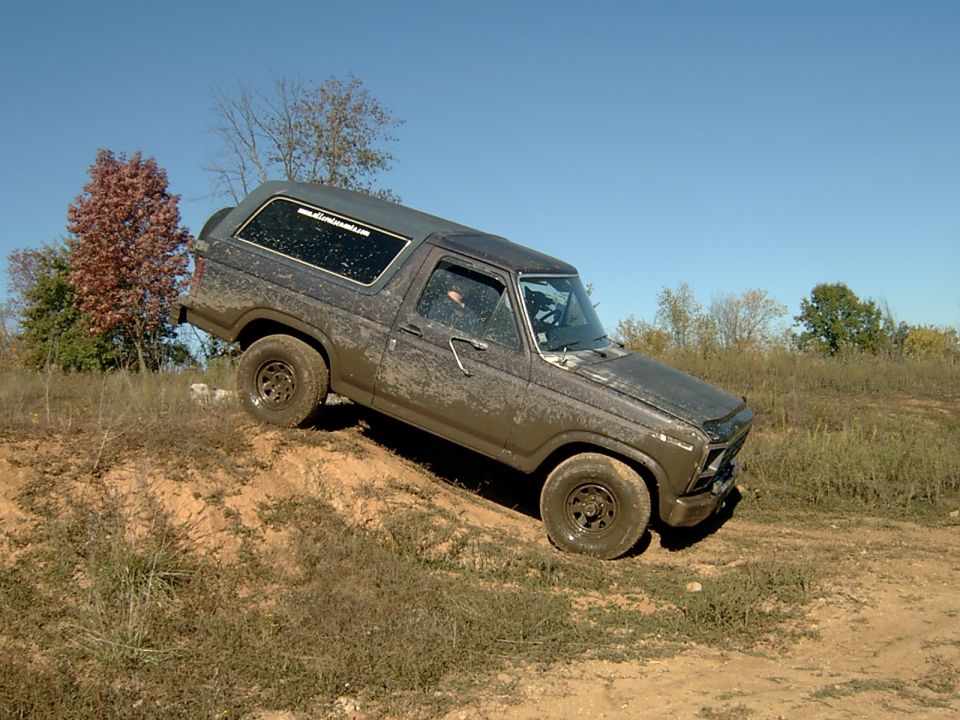 Muddy full-size Ford Bronco driving down a dirt bank in a field, driver's side view